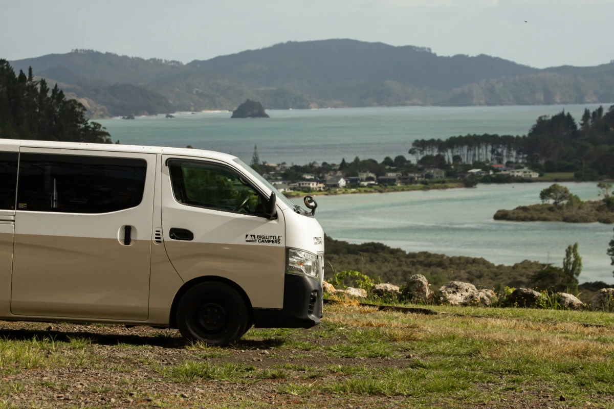 Big Little Campers van parked on a grassy hilltop above a New Zealand bay in summer