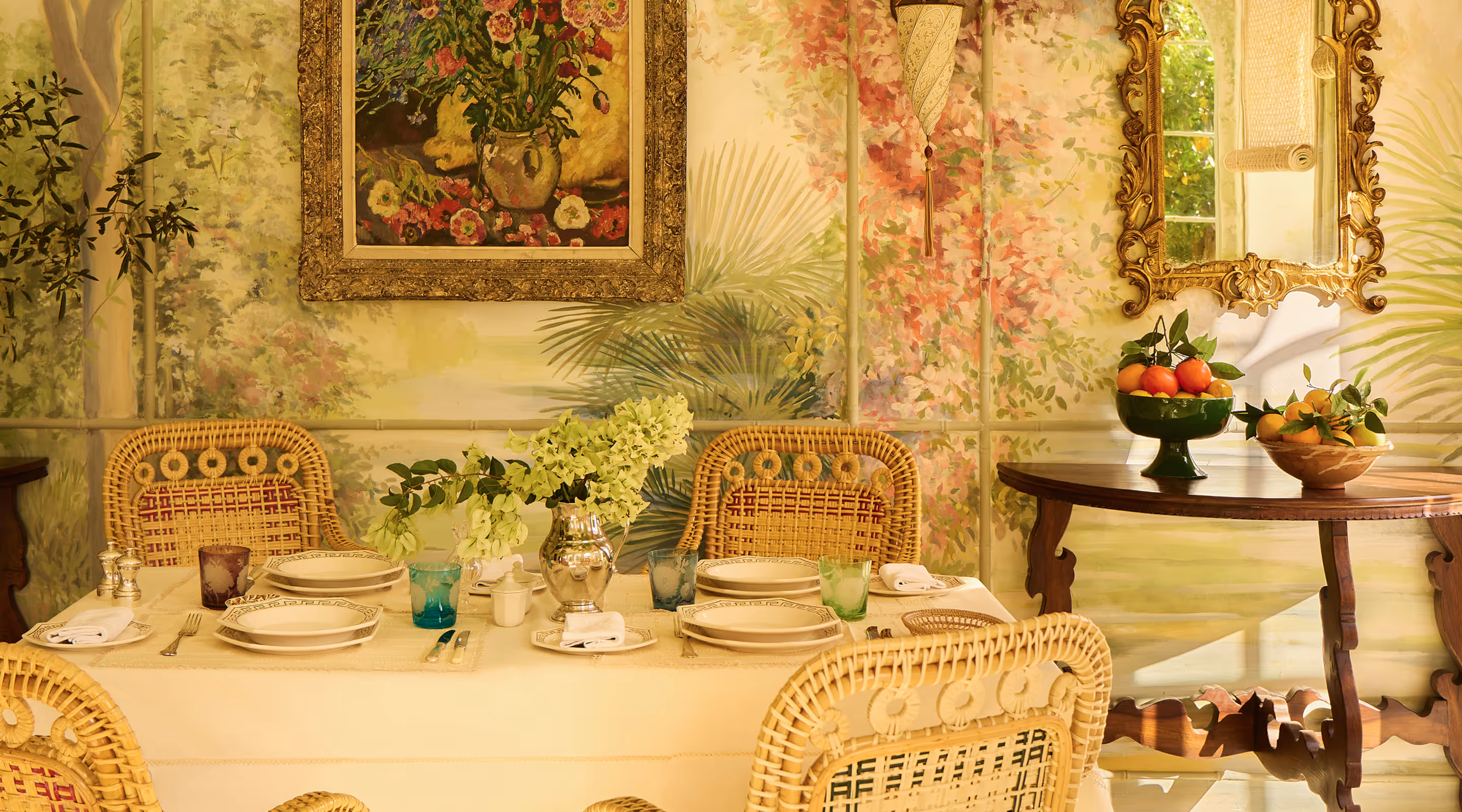 Elegant dining table set with patterned plates, colored glasses, white napkins, and a silver vase with green flowers in a room with floral wallpaper and wicker chairs.