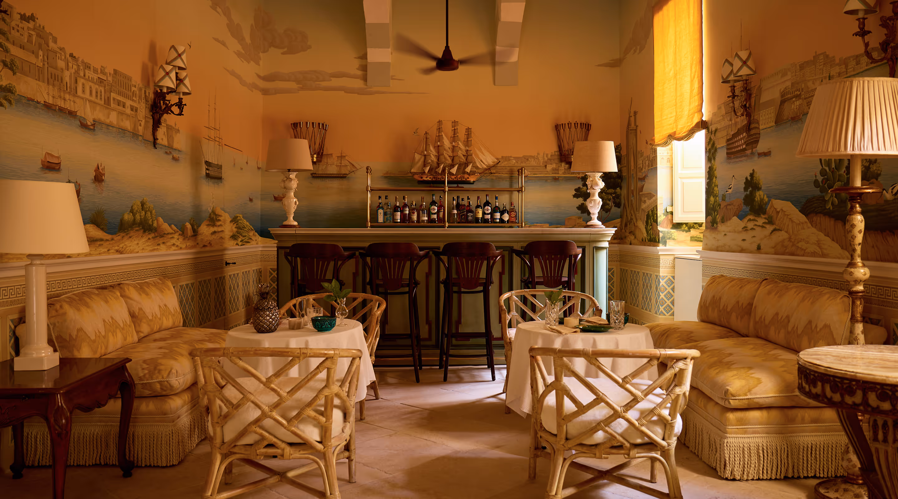 Cozy vintage-style lounge with patterned sofas, wicker chairs, small tables, and a bar counter with bottles and ship decorations.