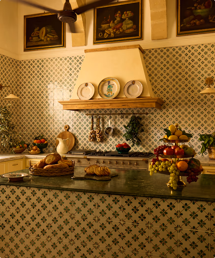 Traditional kitchen with patterned green and white tiles, a stove with hanging garlic and herbs, decorative plates, bread basket, and a tiered fruit stand on a green countertop.