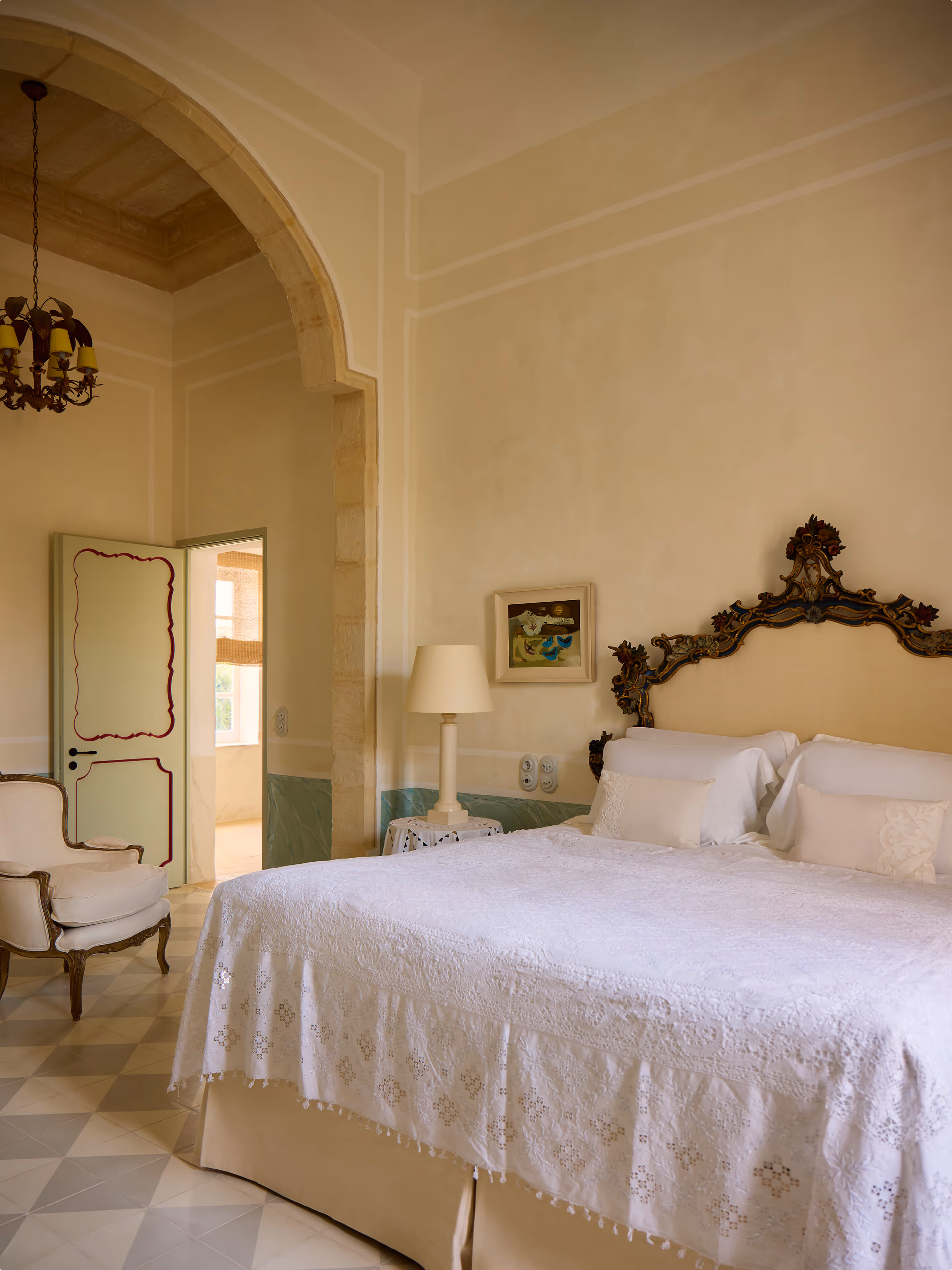 Elegant bedroom with ornate headboard, white lace bedspread, beige armchair, and arched doorway.