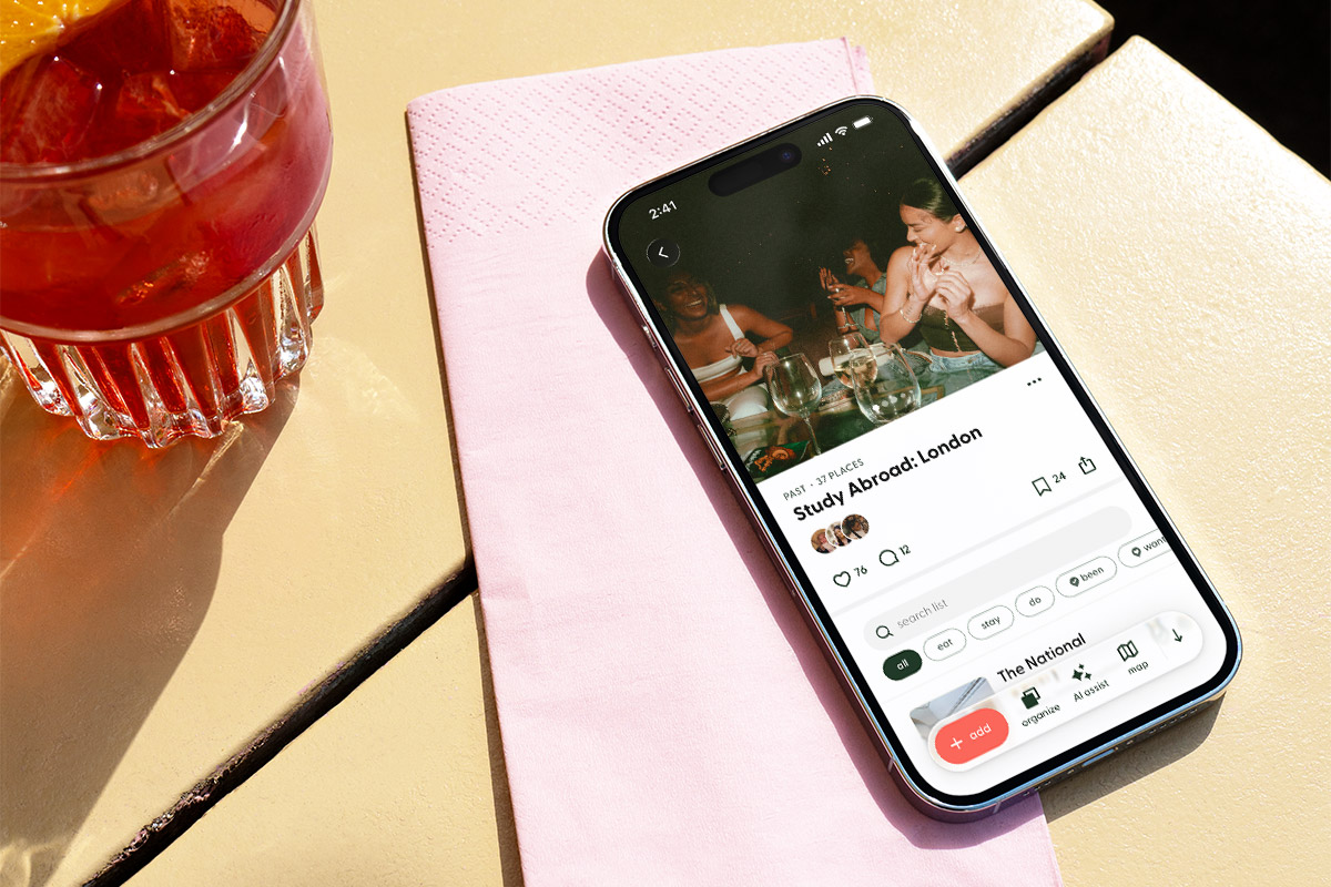 A smartphone rests diagonally on a folded pink napkin atop a cream-colored café table beside a glass of red-orange cocktail garnished with an orange slice. The phone displays a page from the Flaire mobile app featuring a social collection titled “Study Abroad: London,” with a photo of friends gathered around a table, engagement icons, and navigation controls visible on the screen. Bright sunlight casts strong shadows across the tabletop, creating a warm, lifestyle-focused scene.