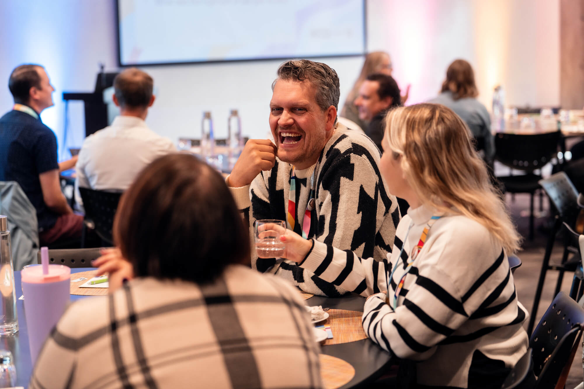 Group of people engaged in conversation at a social event, with a man laughing and a woman holding a glass of water.
