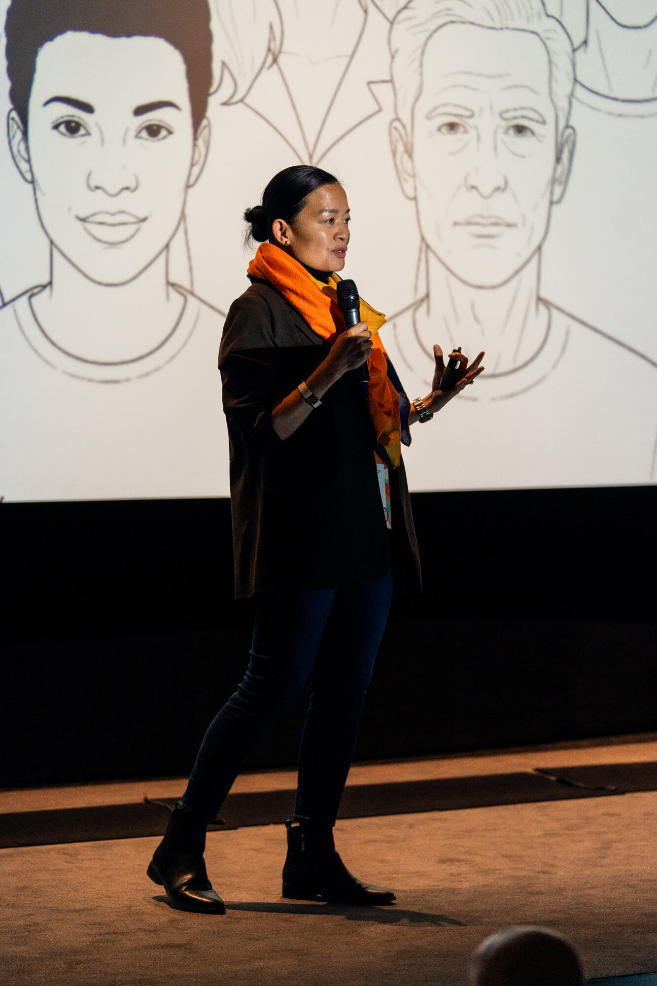 A woman wearing a brown jacket and orange scarf speaking into a microphone in front of a screen with black and white illustrated faces.