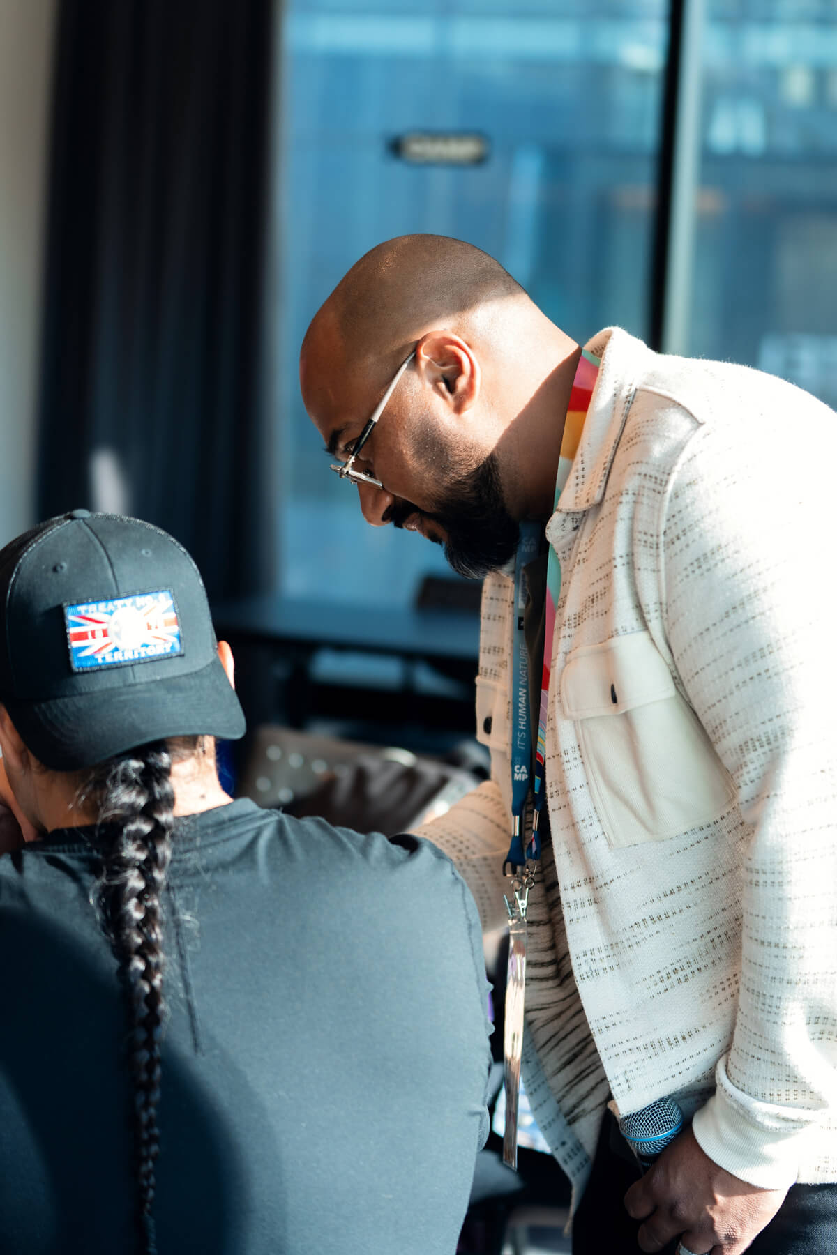 Man with glasses and beard leaning over and interacting with a person with a long braid wearing a black cap and shirt.