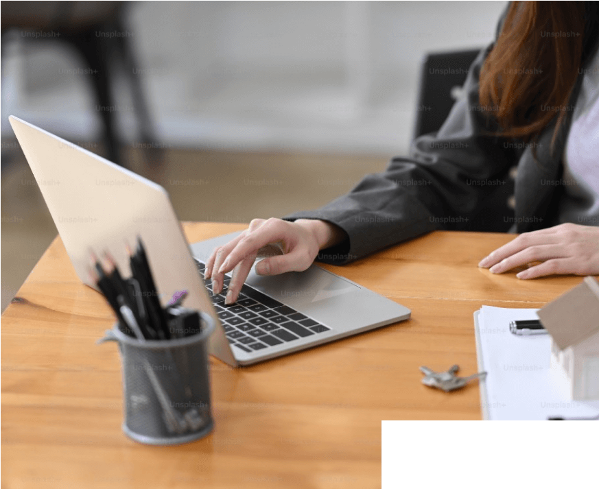 A woman sitting at a desk with a laptop, with a house visible in the background.

