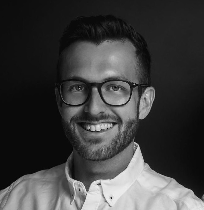 A black and white photo of a man wearing glasses, looking thoughtfully off to the side.


