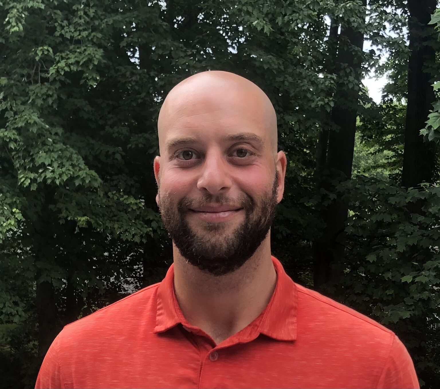 Smiling bald man with a beard wearing an orange collared shirt standing outdoors with green leafy trees in the background.