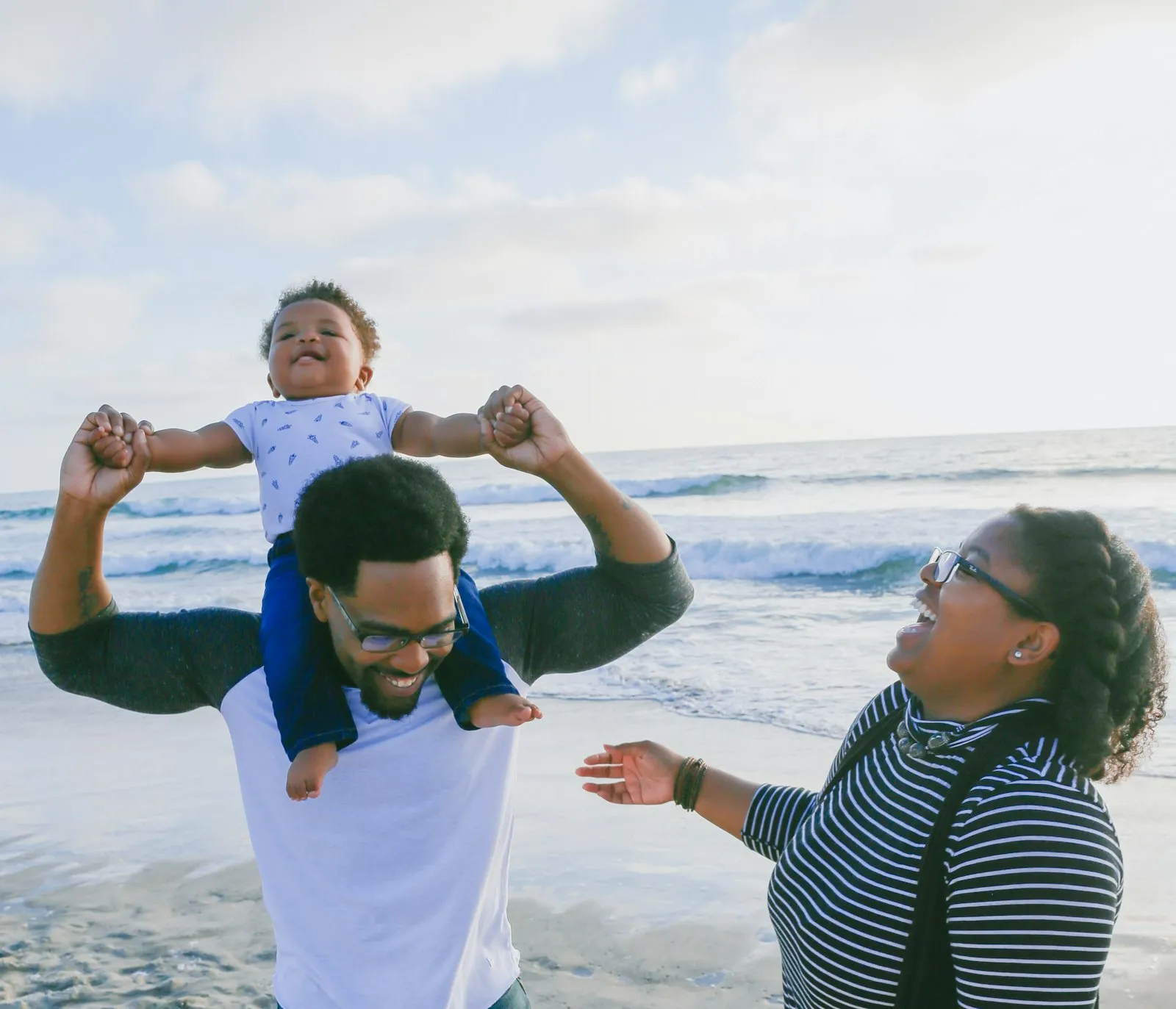 A family laughing together at the beach as a father lifts his smiling baby onto his shoulders.