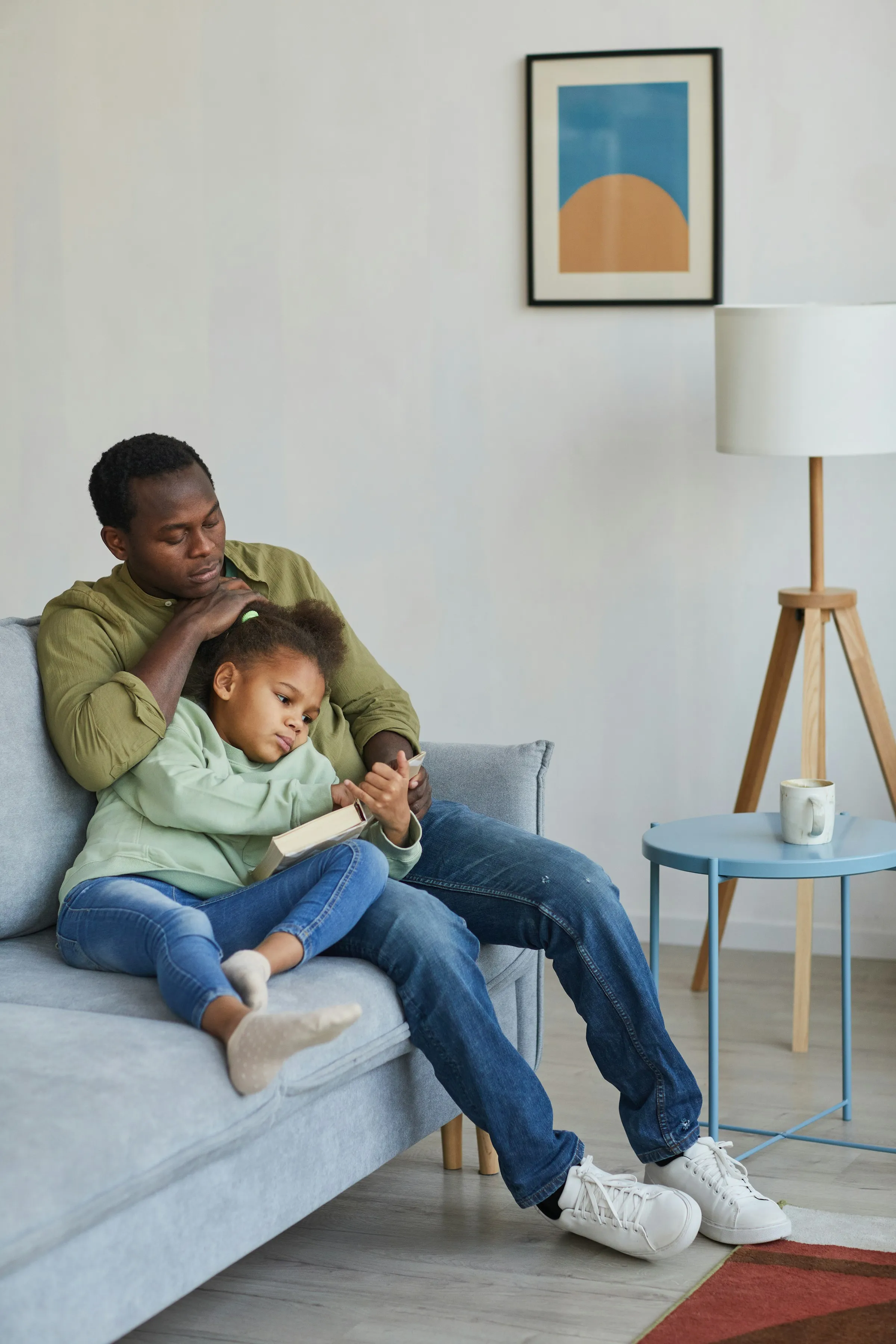 A father and his daughter read together on the living room sofa.