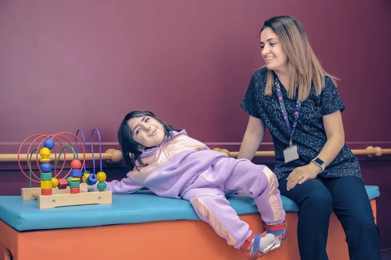 A behavior analyst smiling beside a child playing with a bead maze on a therapy mat.