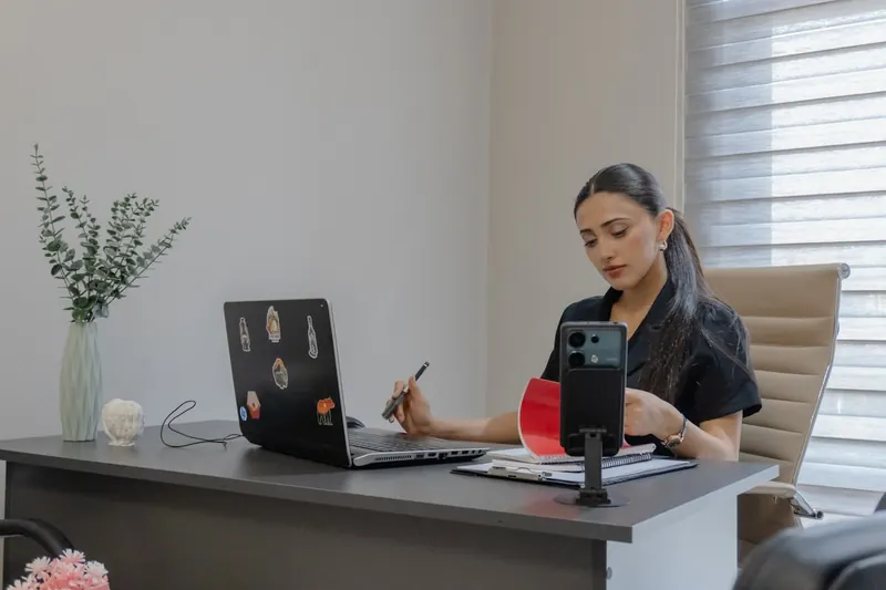 A person reviewing paperwork at a desk with a laptop and phone.