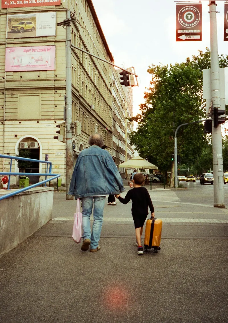 A grandfather and a young child walking together, the child pulling a small suitcase.