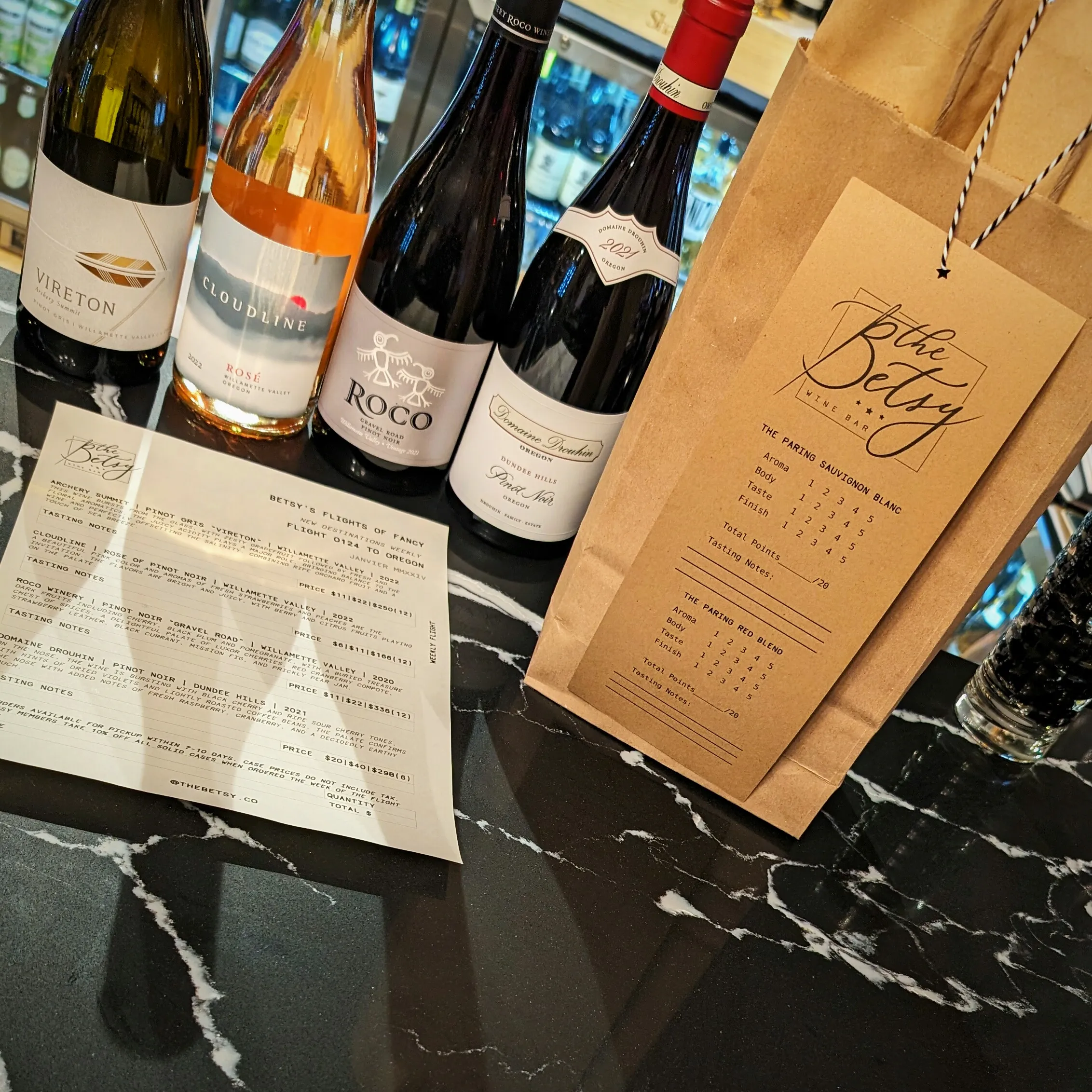 Photo is of four bottles of curated wines that members of The Betsy get to taste weekly as part of their membership. Plus a bag that holds additional wines they get to leave in their locker or bring home with them. 
