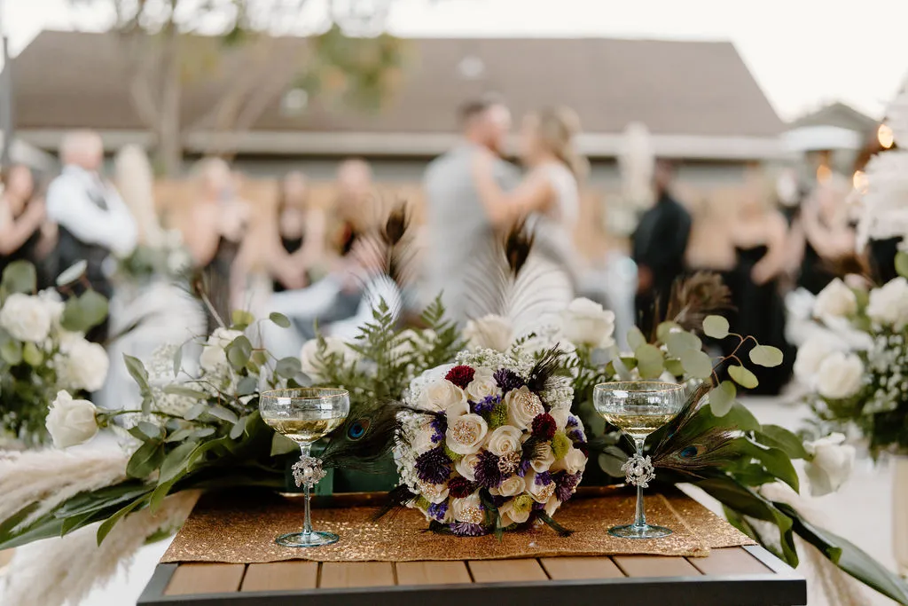Beautiful floral boutique and centerpieces with two wine glasses in the foreground with the bride and groom dancing in the background as their friends and family watch. Event was hosted at The Betsy.