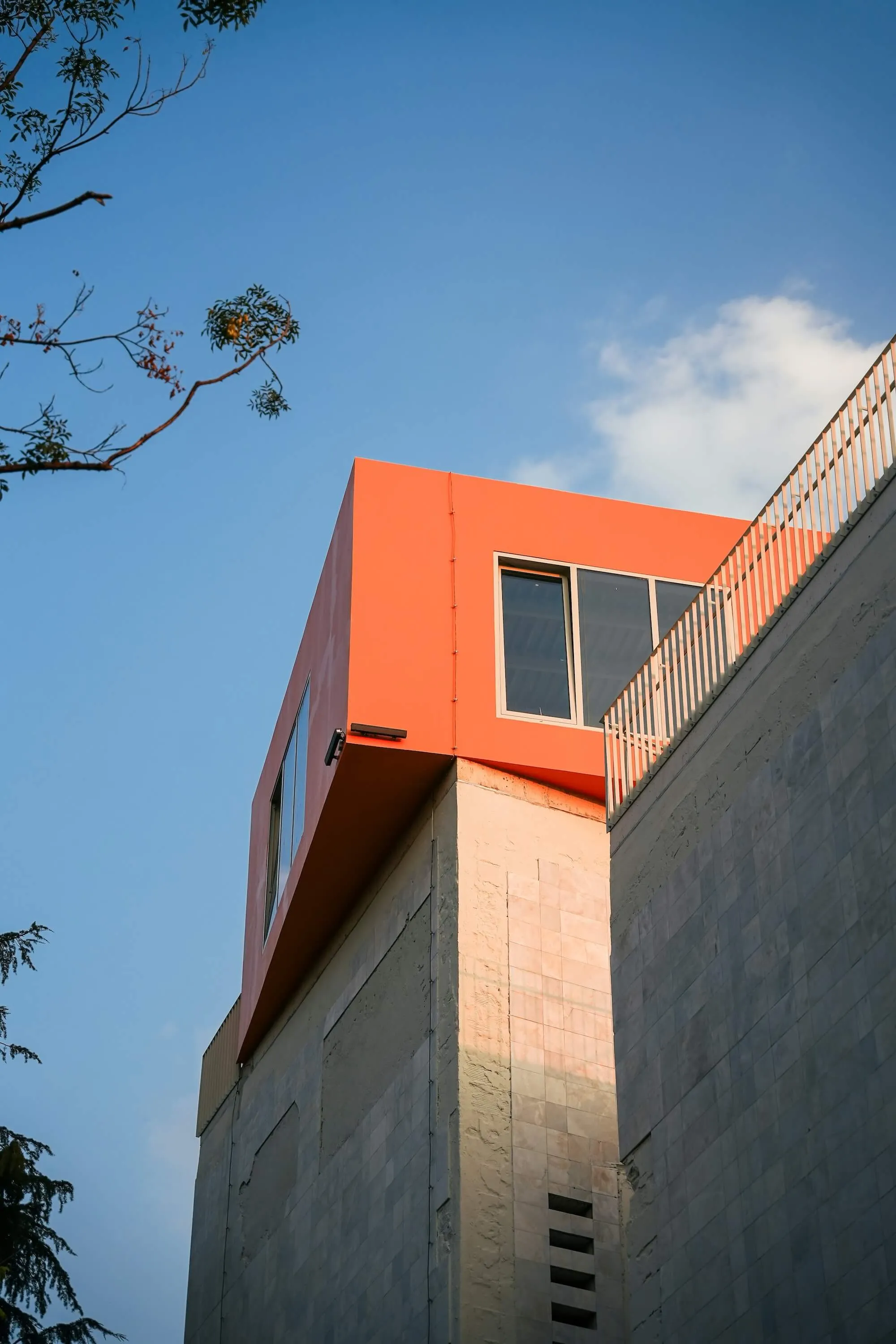 Modern building corner with an orange upper section featuring large windows against a blue sky with some tree branches.