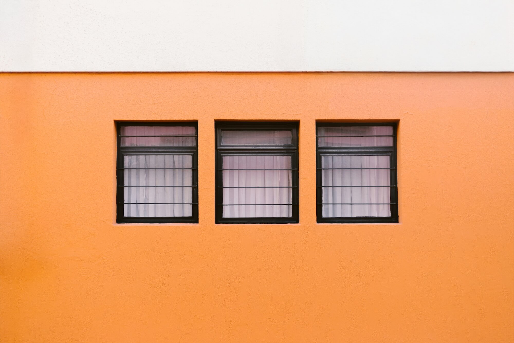 Three black-framed windows with bars and pink curtains set in an orange and white painted wall.