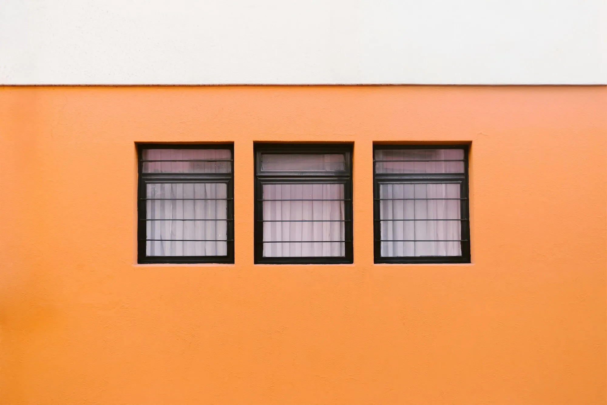 Three black-framed windows with bars and pink curtains set in an orange and white painted wall.