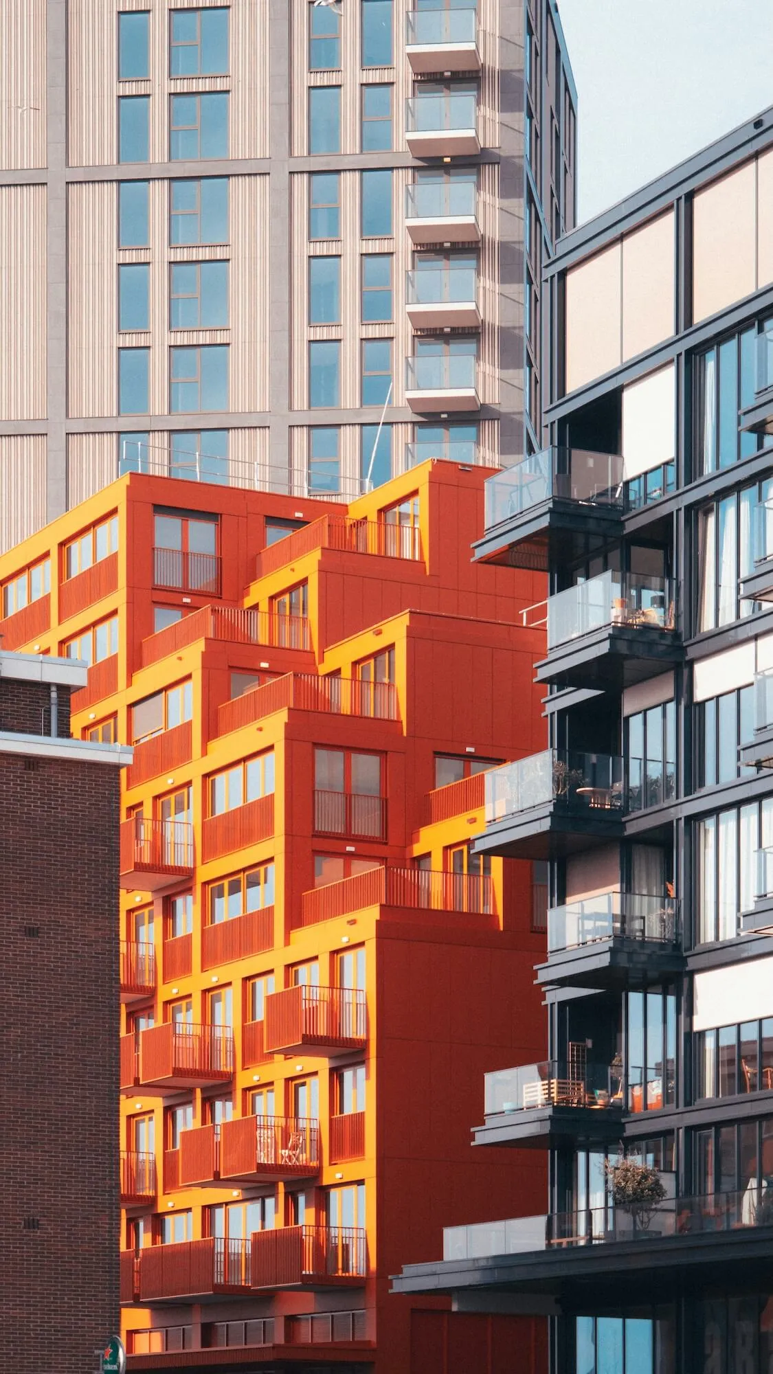 Modern building corner with an orange upper section featuring large windows against a blue sky with some tree branches.