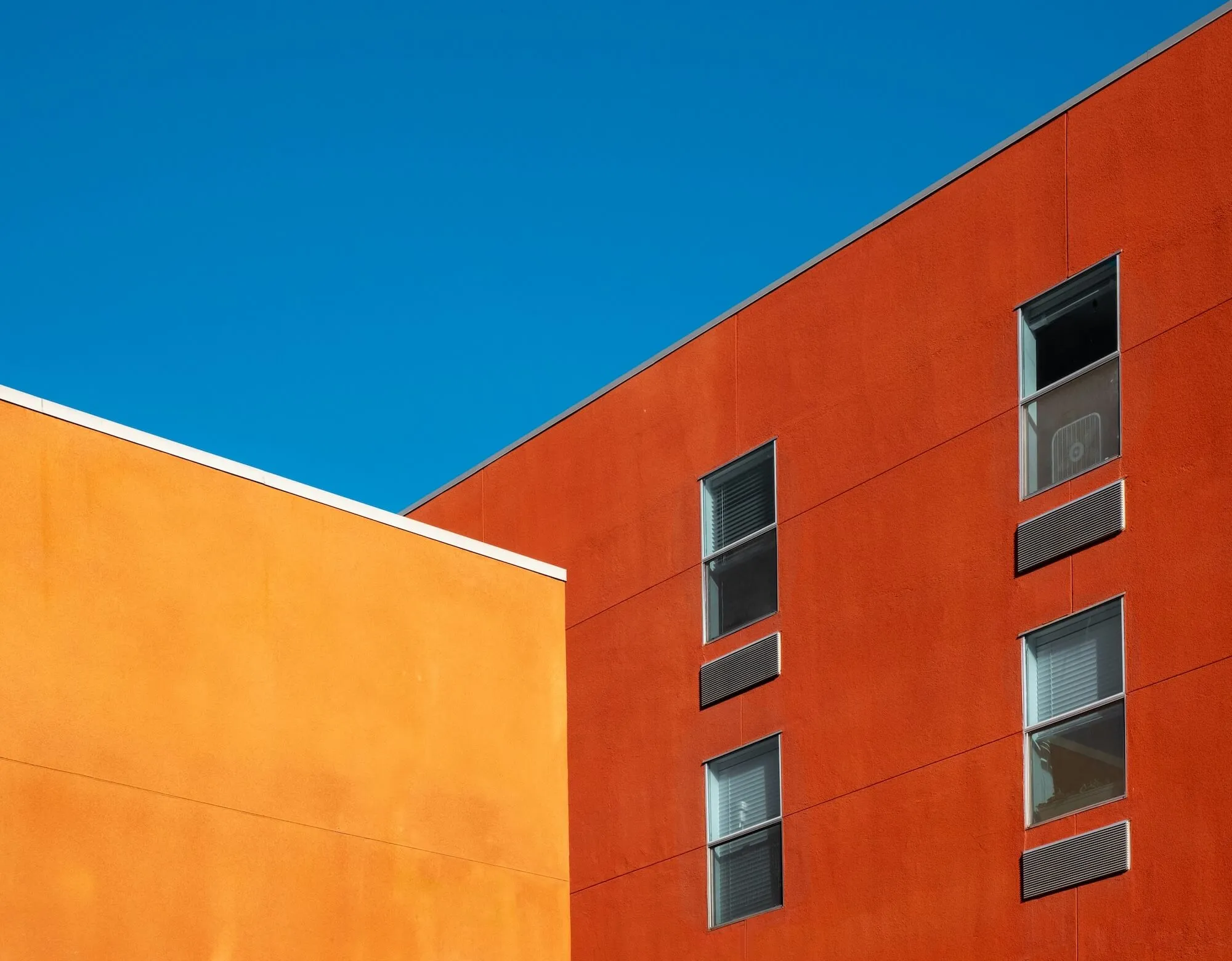 Modern building corner with an orange upper section featuring large windows against a blue sky with some tree branches.