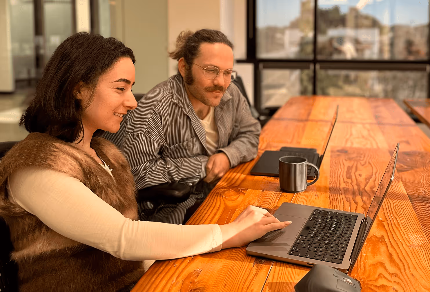 Two people sitting at a wooden table collaborating while looking at a laptop screen.