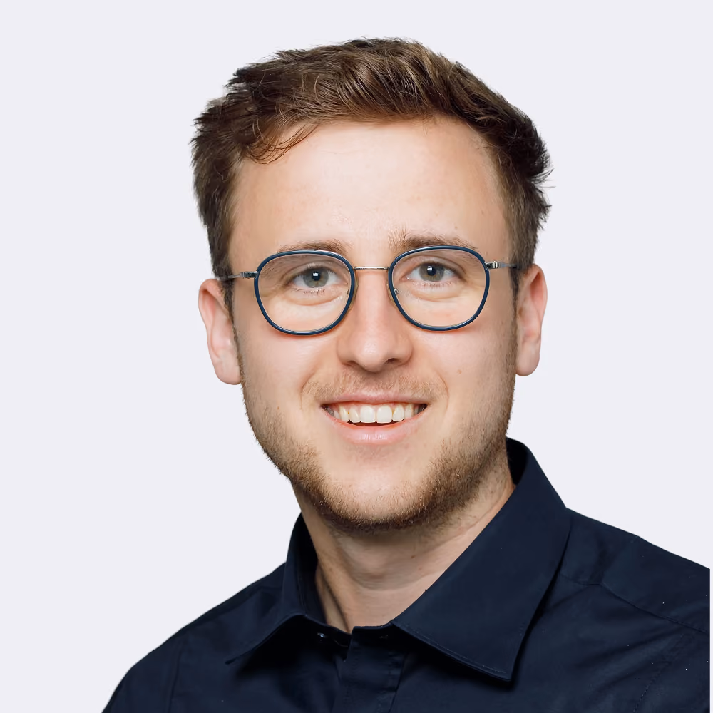 Junger Mann mit kurzen braunen Haaren, Brille und dunkelblauem Hemd lächelt vor hellem Hintergrund.