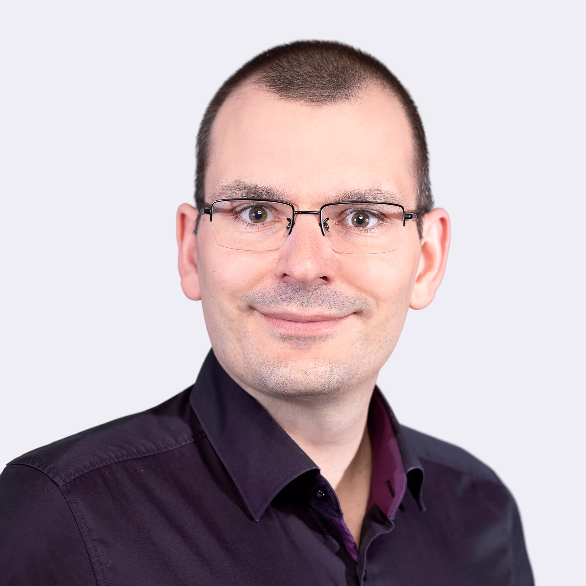 Lächelnder Mann mit Kurzhaarschnitt und Brille vor hellem Hintergrund.