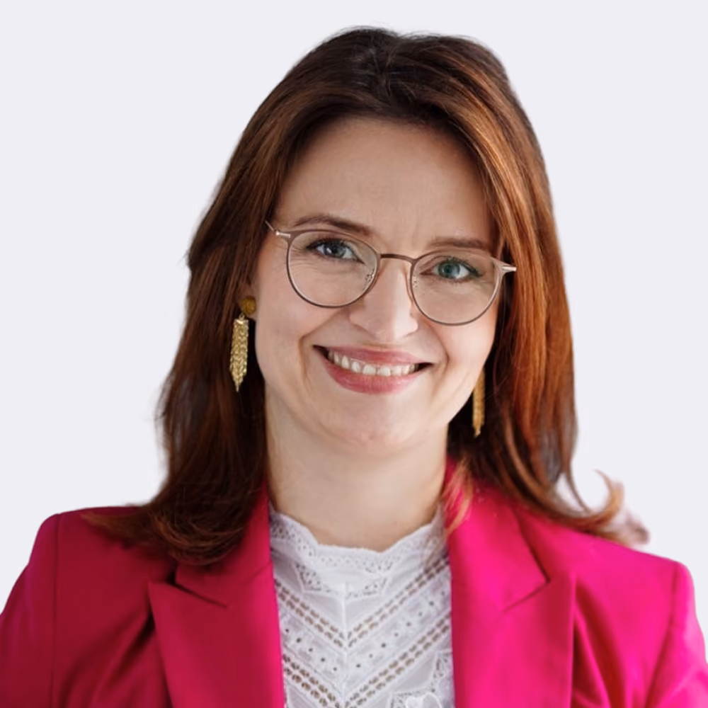 Frau mit roten Haaren, Brille, goldenen Ohrringen und pinkem Blazer lächelt freundlich.