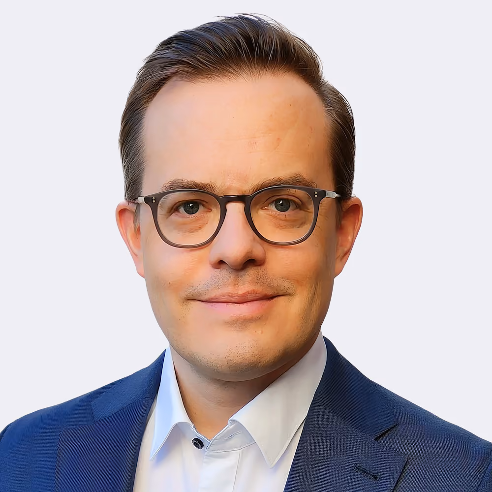 Mann mit kurzen braunen Haaren, Brille und blauem Anzug lächelt vor hellem Hintergrund.
