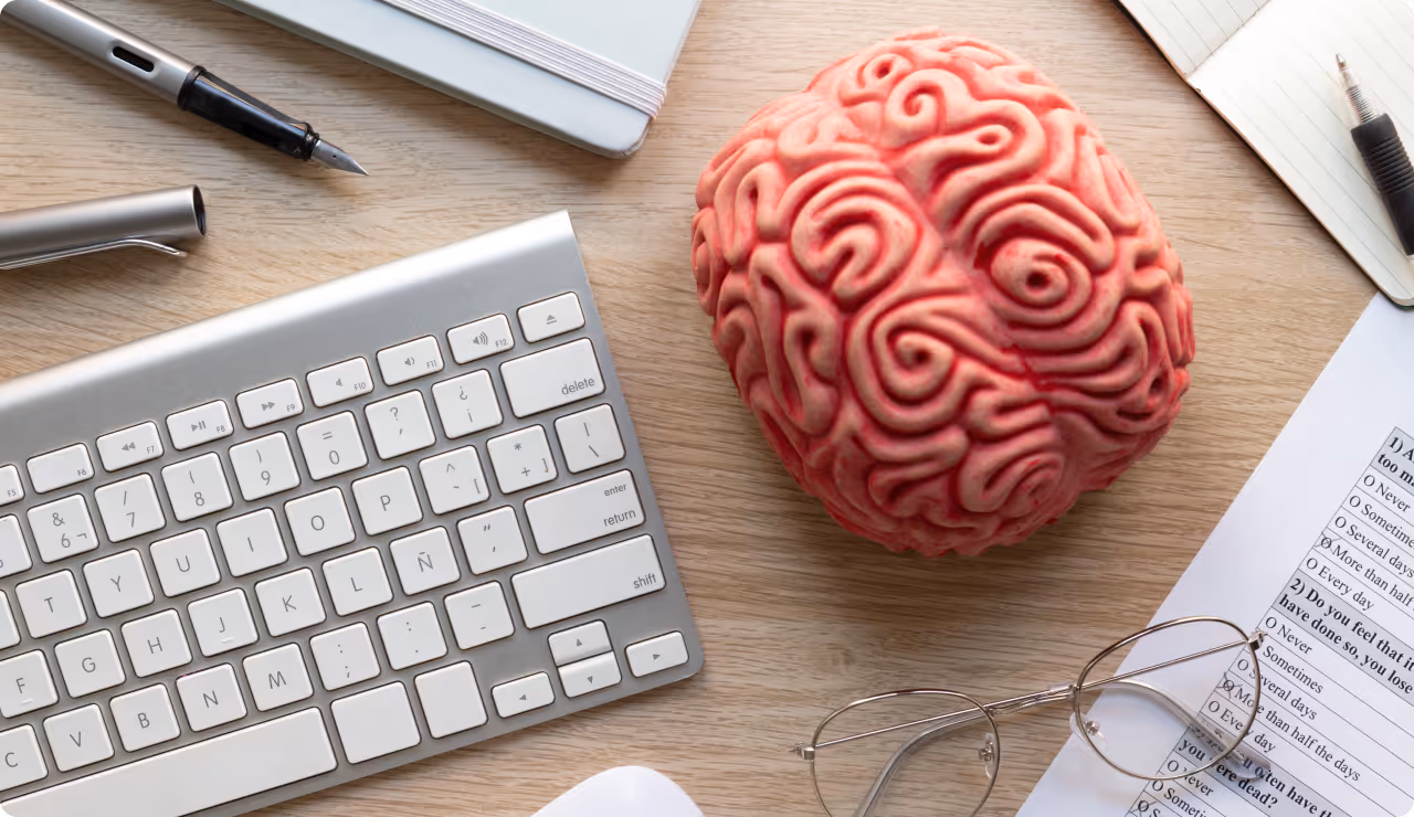 Gros plan sur un cerveau rose stylisé, posé sur un bureau à côté d’un clavier et de lunettes, symbolisant le fonctionnement cognitif.