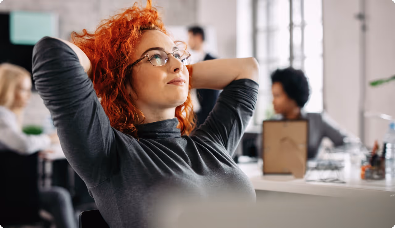 Femme rousse assise en arrière sur sa chaise, confiante et détendue dans un open-space.