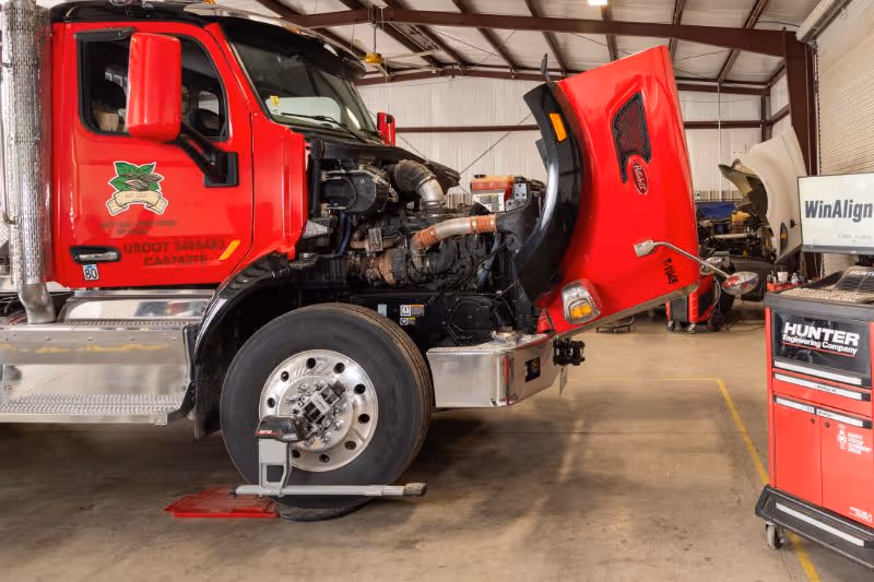 A white semi-truck is parked in a clean repair bay with its hood open. Diagnostic equipment stands nearby, and shop tools and blue lifts are visible in the background. 
