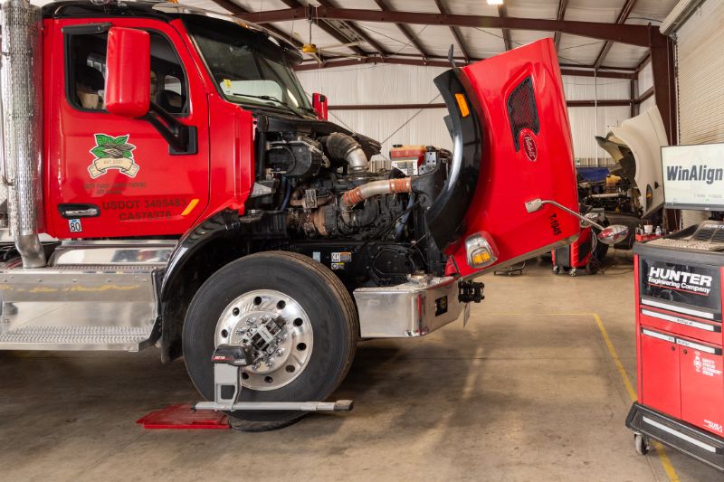 A white semi-truck is parked in a clean repair bay with its hood open. Diagnostic equipment stands nearby, and shop tools and blue lifts are visible in the background. 