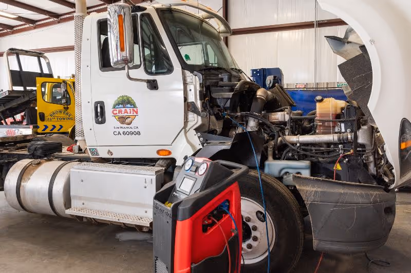 White semi-truck with hood open in repair shop, diagnostic equipment connected to engine, yellow tow truck and tools visible in background. 