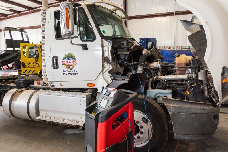 White semi-truck with hood open in repair shop, diagnostic equipment connected to engine, yellow tow truck and tools visible in background. 