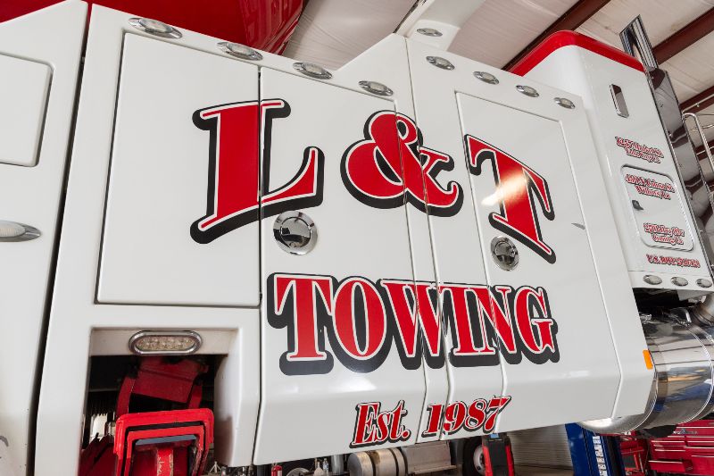 Close-up of a heavy-duty tow truck displaying bold red L & T Towing graphics and Est. 1987 lettering on white body.