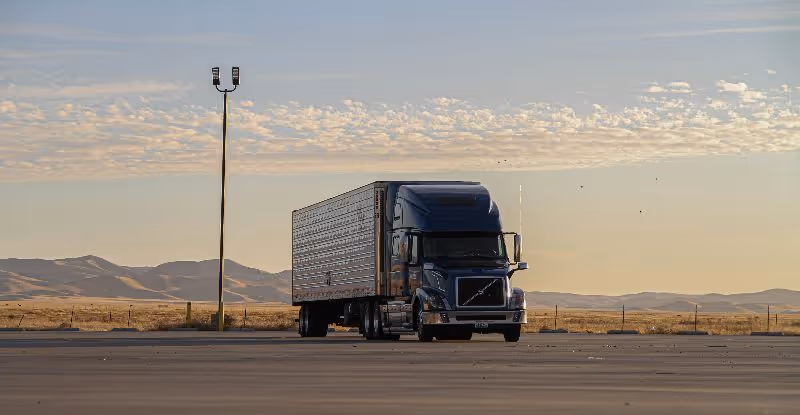 Black Volvo semi truck parked in open lot with desert mountains and cloudy sky at sunset background.