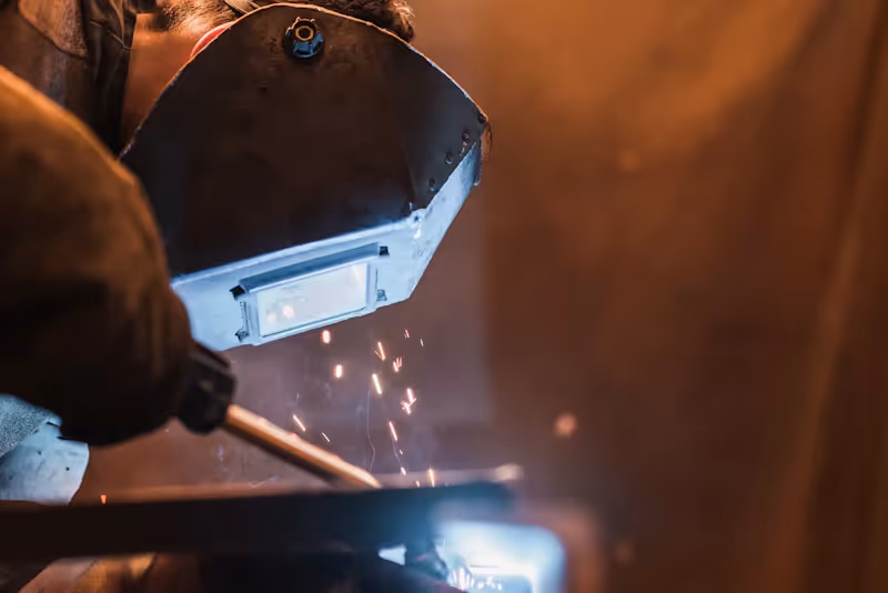 Trailer fabrication service showing technician welding heavy-duty metal component with sparks and protective welding shield.