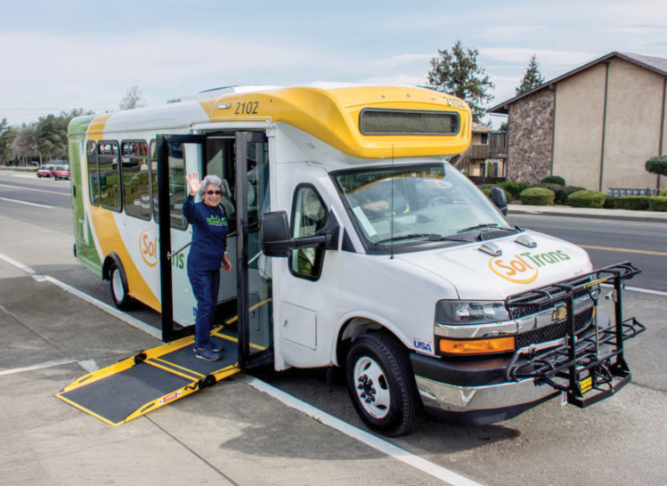 A white and yellow SolTrans shuttle bus is stopped on the side of the road with its front wheelchair ramp extended. A woman wearing sunglasses and a navy blue outfit is standing at the open bus door waving. The bus has a bike rack attached to its front and is parked near residential buildings with trees in the background.