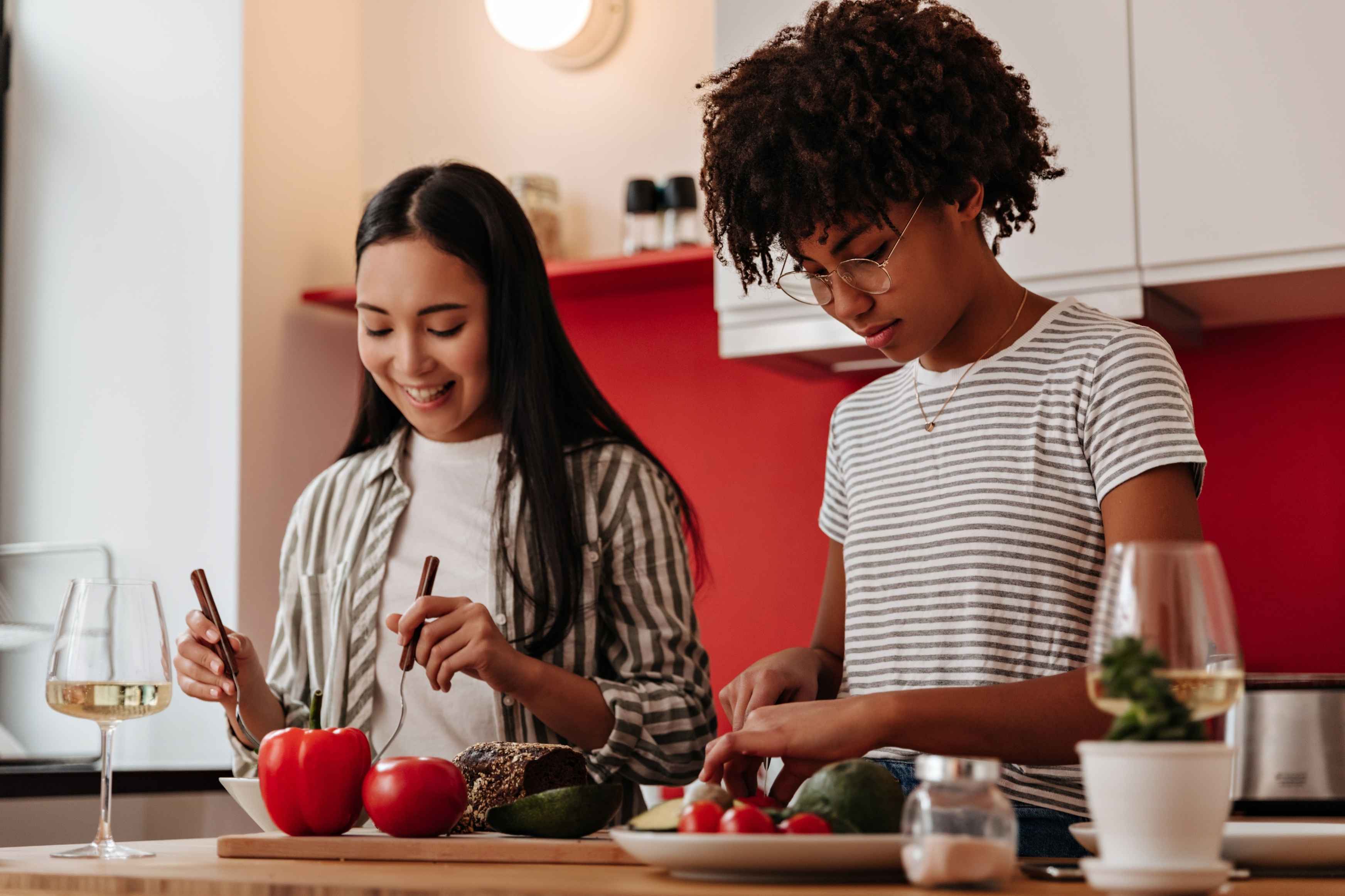 Two young women preparing food together in a modern kitchen one slicing vegetables while the other smiles, holding cutlery near fresh produce on the counter. 