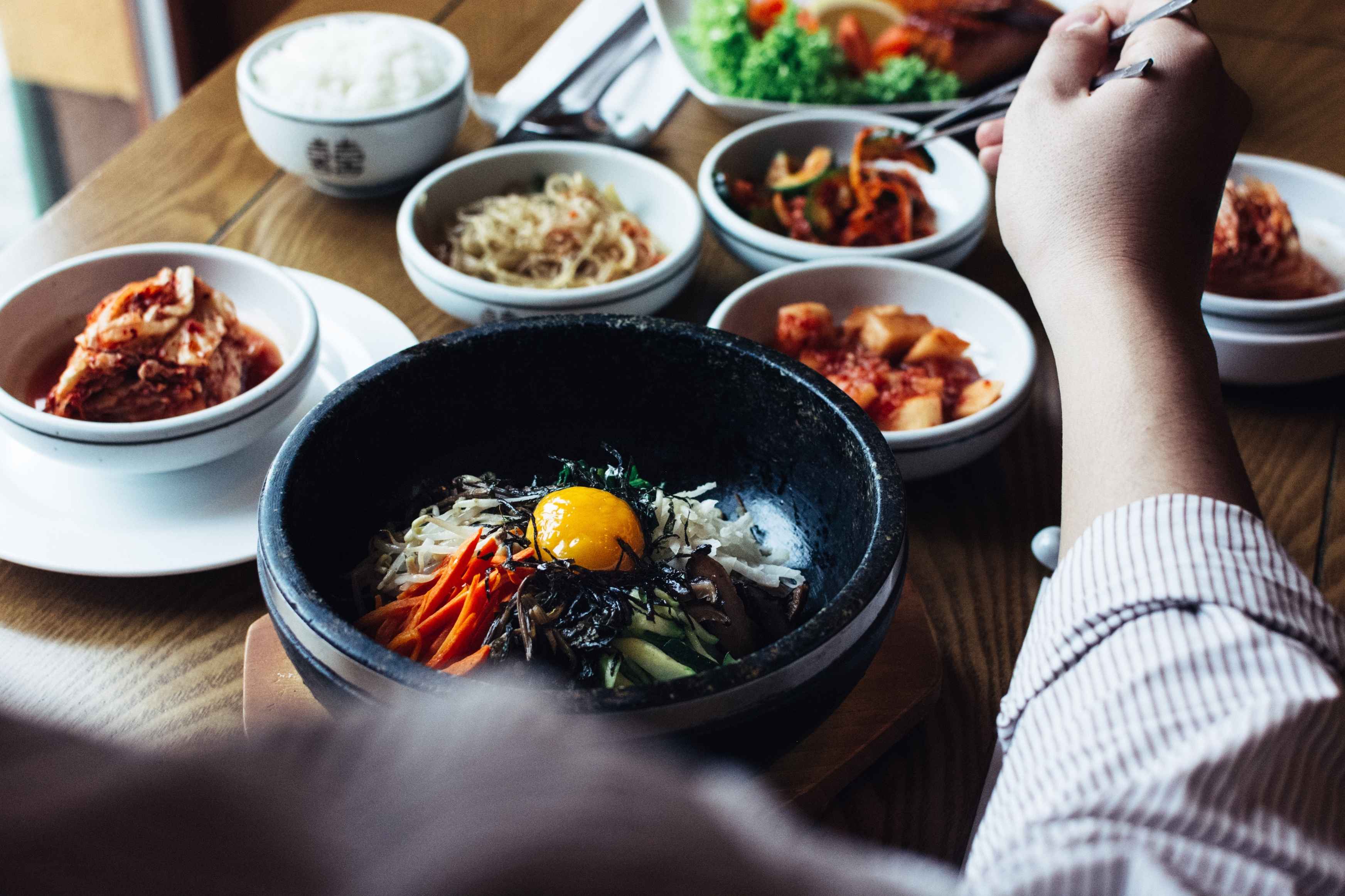 A person enjoys a Korean meal with bibimbap in a stone bowl and assorted vegetable side dishes.