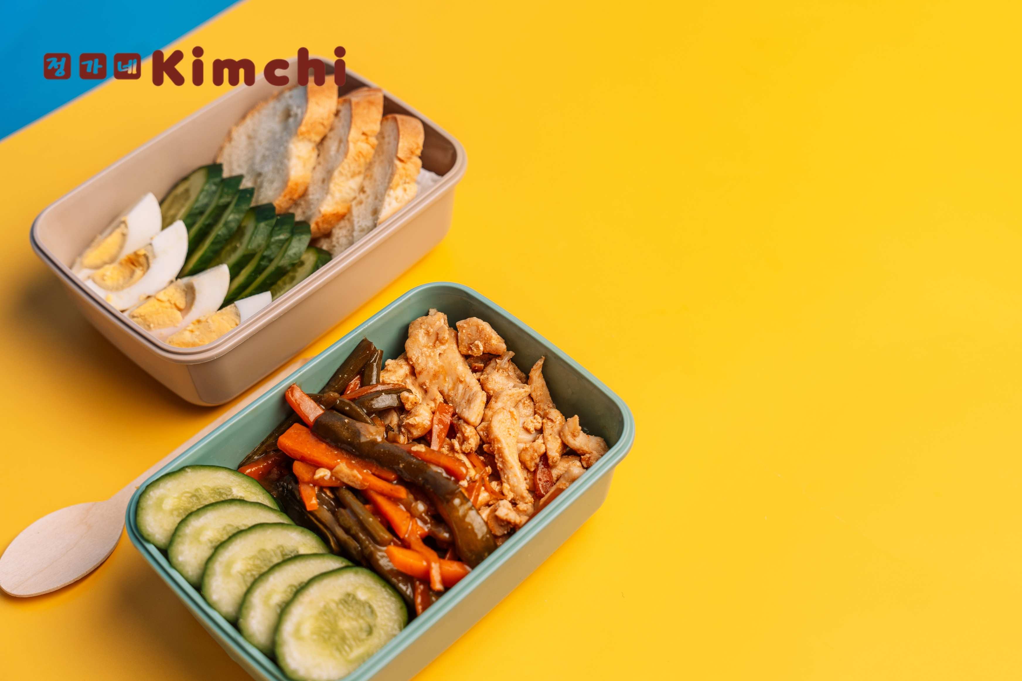 Two colourful bento boxes on a vibrant yellow background. One box contains sliced hard-boiled eggs, cucumber, and toasted bread; the other features Korean BBQ chicken, stir-fried vegetables, and cucumber slices.