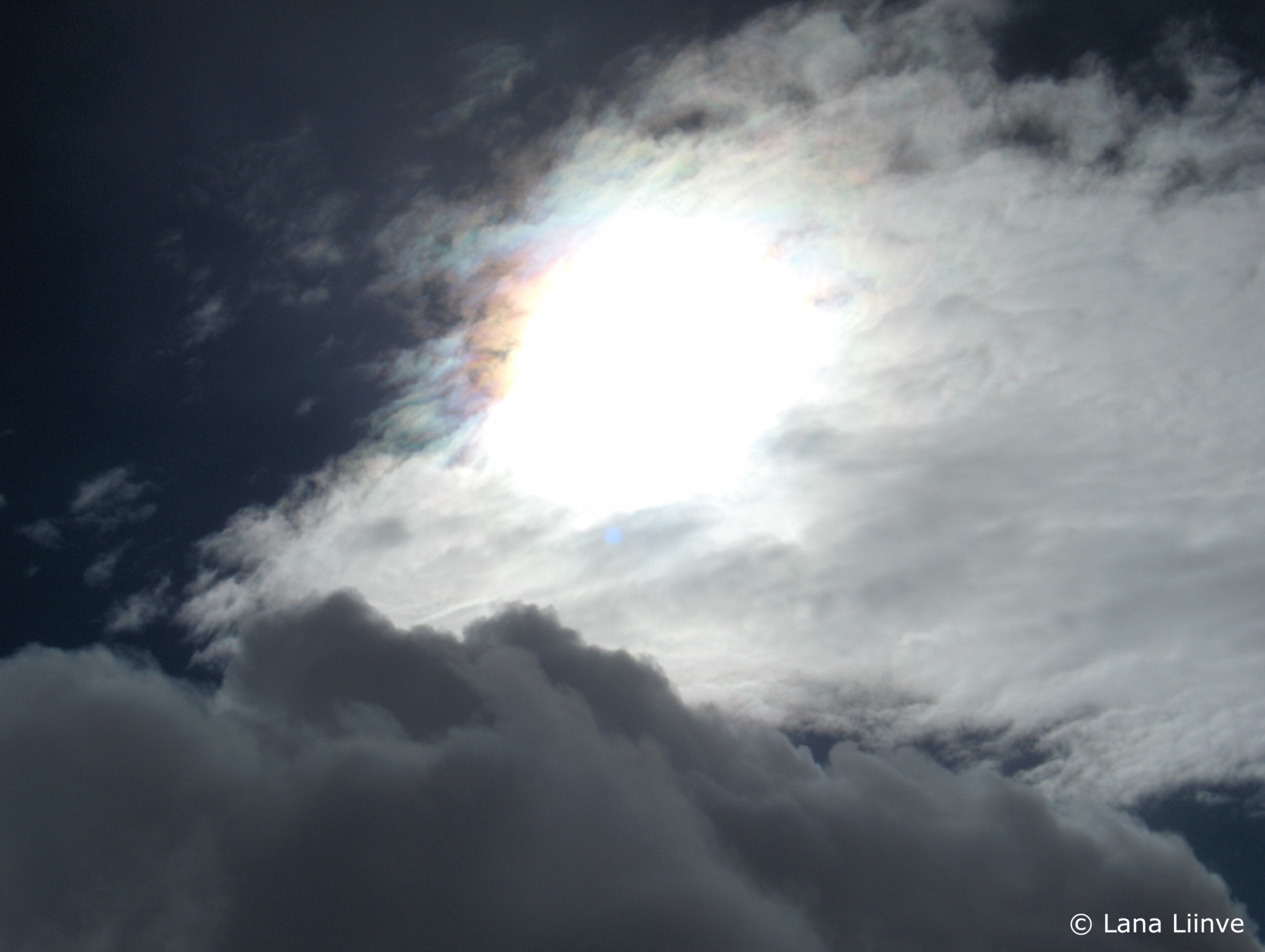 A photo taken directly at the sun shining bright behind some thick white clouds, where a rainbow-colored ring presents partially around the edge of the sun, and a dark blue sky is present in the background