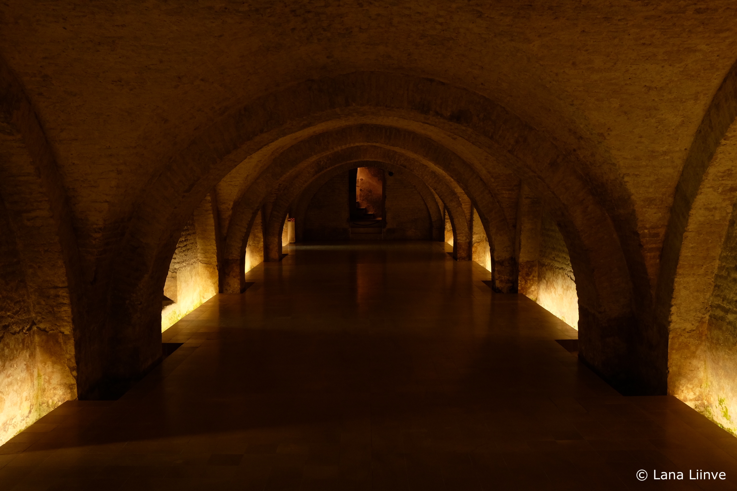 A photo showing an underground multi-arched chamber lit by golden lights, with a small staircase with orange-hued light visible in the central background, at the Real Alcázar in Sevilla, Spain