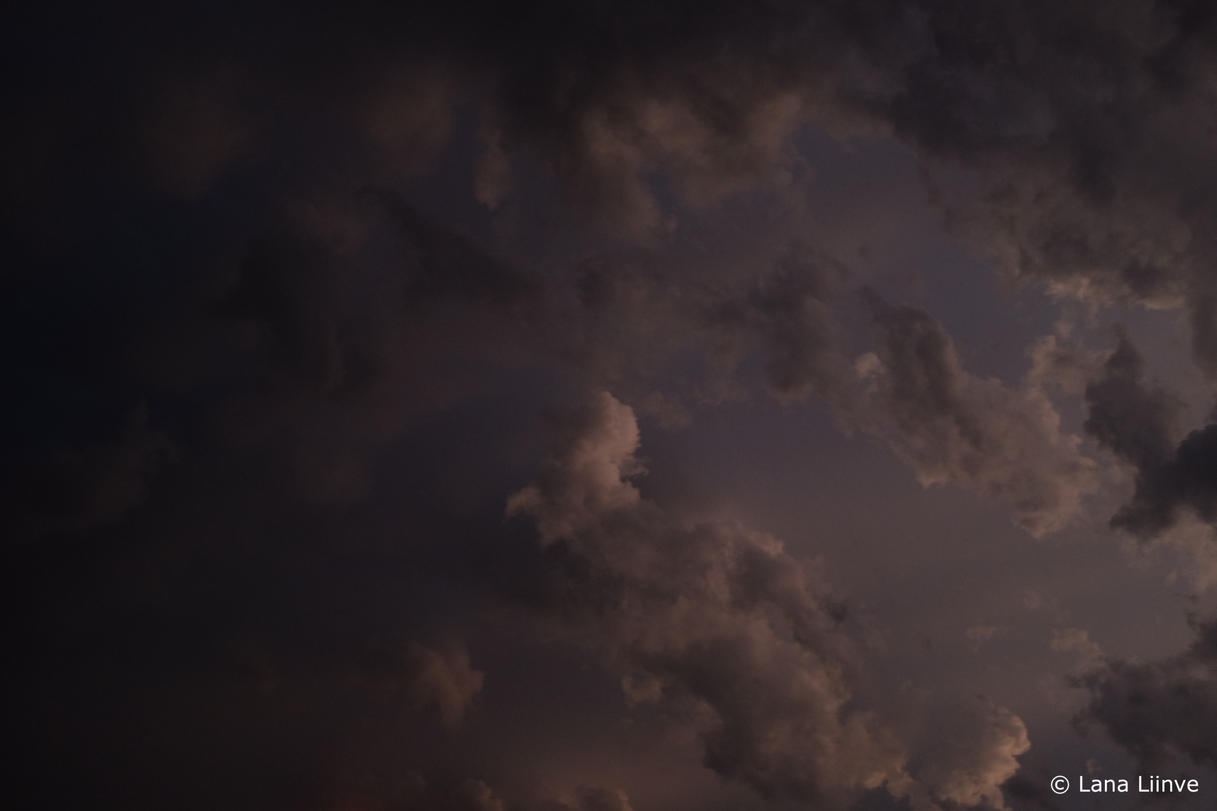 A photo taken looking into a dark,stormy late-afternoon sky, where in the last remaining light, fluffy clouds are barely visible in off-white, grey, and purple hues