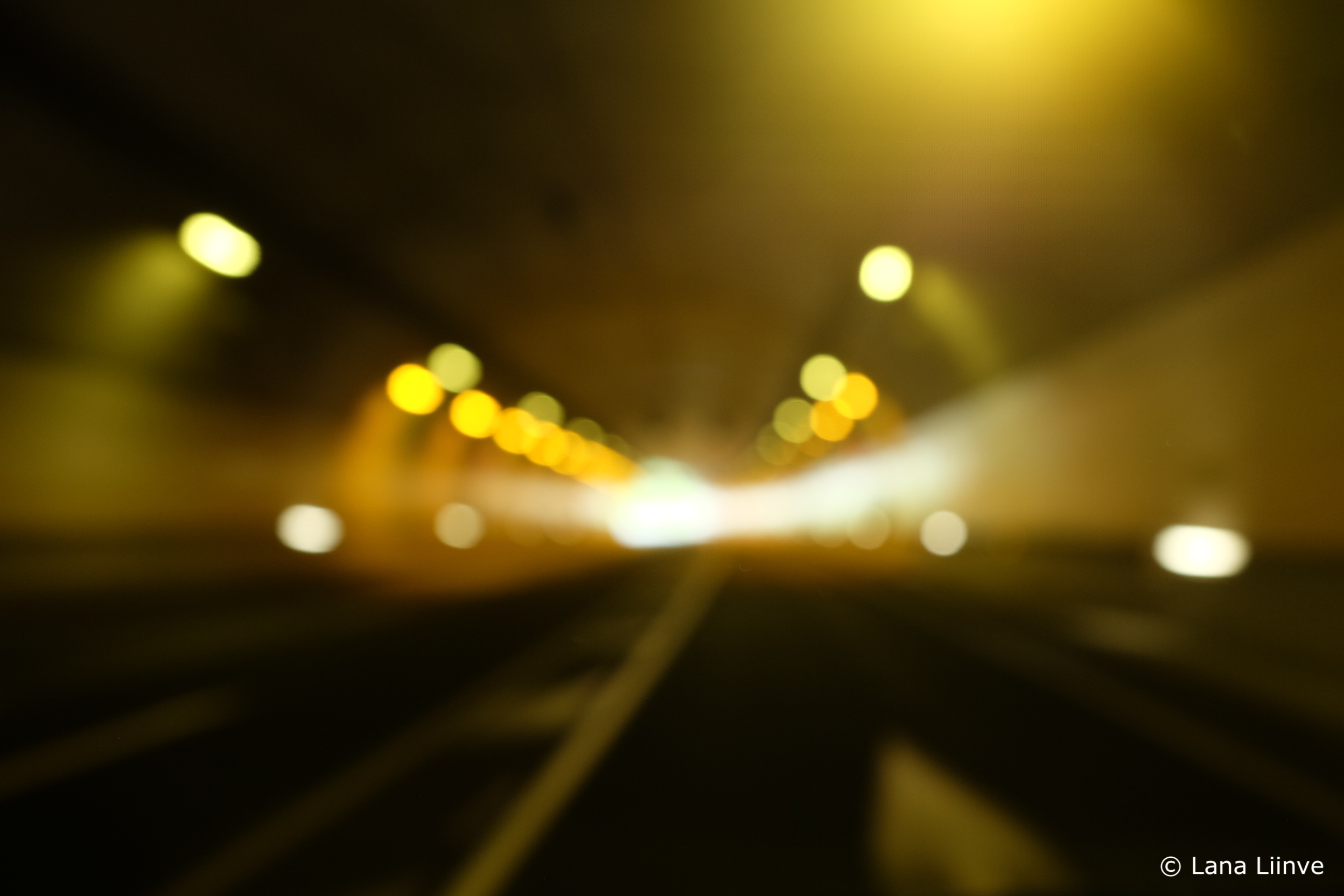 A blurry photo taken while driving through a tunnel, showing dark pavement with white lines, bright golden yellow lights scattered about the tunnel, and a bright light-colored opening at the central end