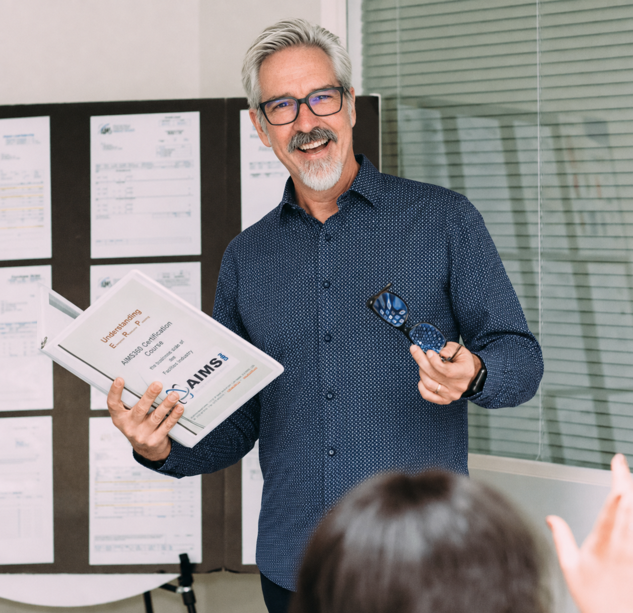 AIMS360 trainer Scott Allen holding the Understanding ERP certification course binder while teaching a class, with a student raising a hand