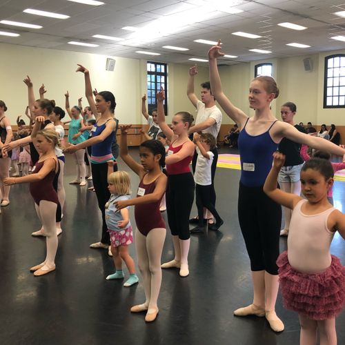 Several dancers with arms raised that vary in ages within a classroom.