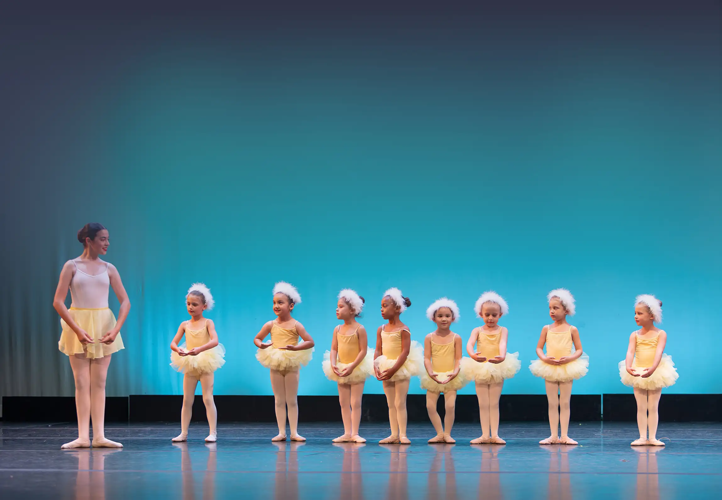 A stage showing a ballet teacher and several children ballet students in yellow
