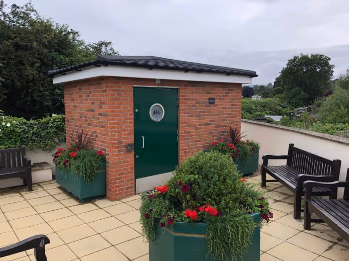 A small brick building with a green door is surrounded by benches and potted plants on a patio.